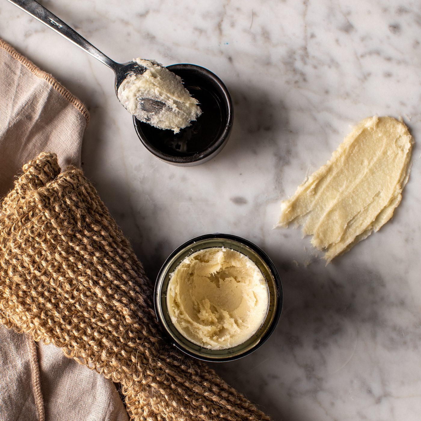 A close-up flat lay of a pale yellow shea butter in two small dark jars on a light marble surface, with a spoon holding some of the product and a textured beige cloth beside it.