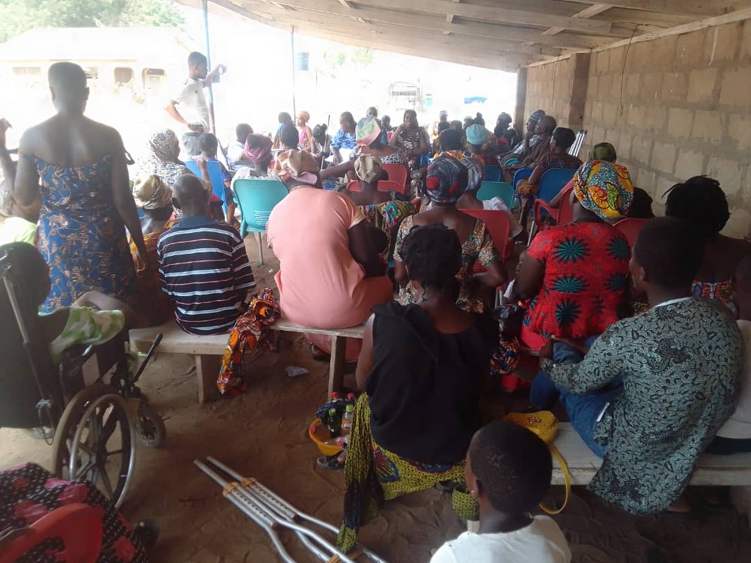 A behind view of large group of Ghanaian people sitting and standing in a semi-open shelter space with concrete walls, chairs, crutches and a wheelchair visible in the foreground.